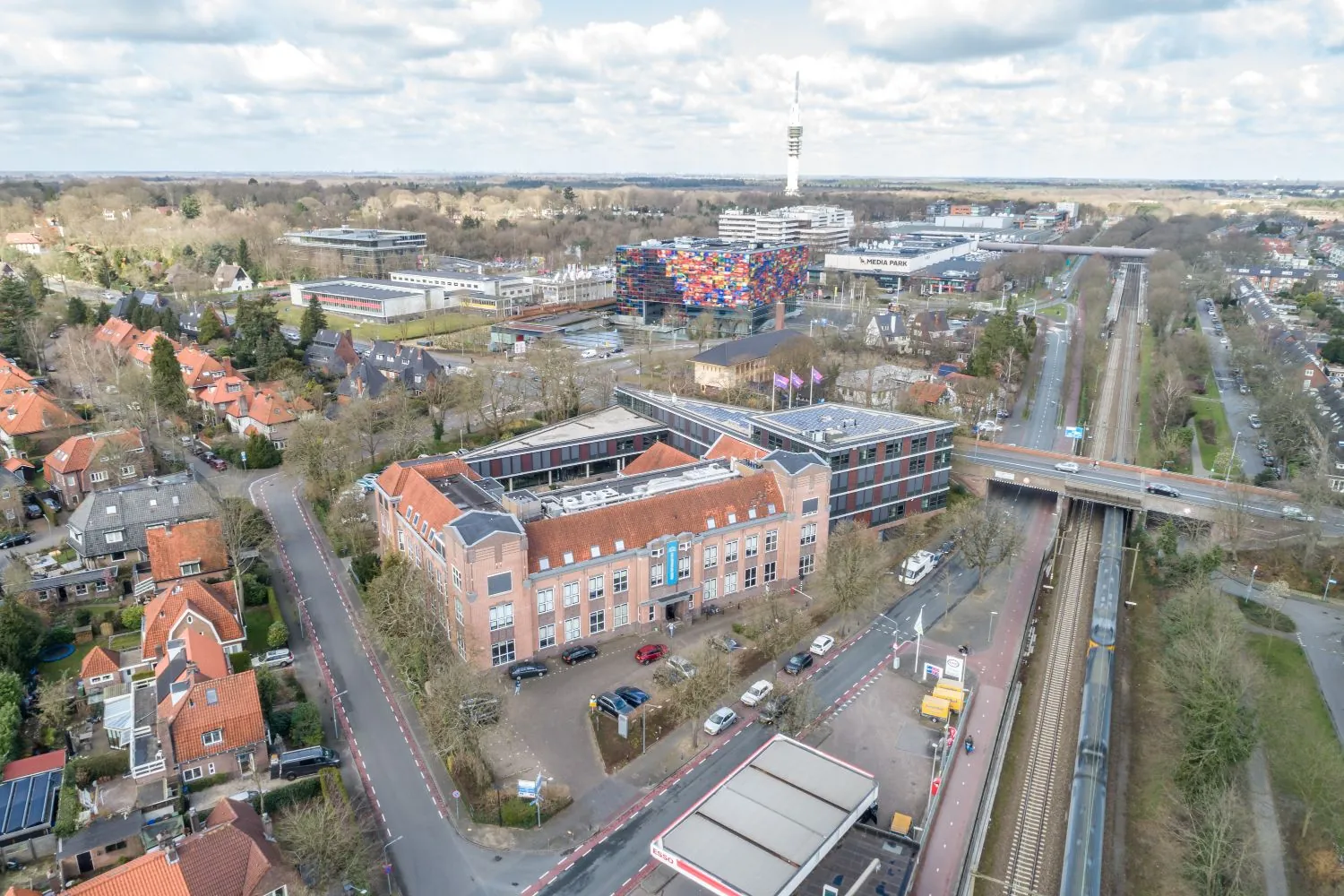 Luchtfoto van de Lage Naarderweg in Hilversum met uitzicht op het Media Park, het kleurrijke Instituut voor Beeld en Geluid en een passerende trein.
